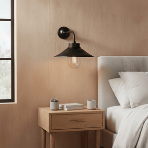 Matt black wall lamp with a cone shade illuminating a bedside table with books and a plant.