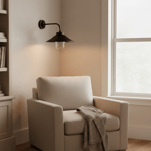 Matt black wall lamp with glass shade illuminating a cozy reading nook beside a beige armchair.