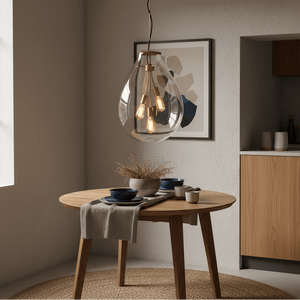 Matt brass pendant light with clear glass shade hanging above a wooden dining table set with dishes.