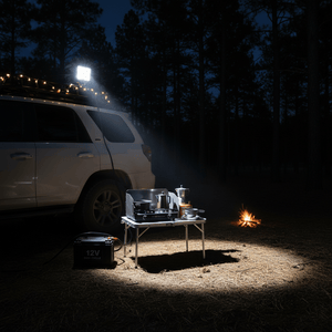 LED work light illuminating a camping setup with a stove and 12V battery at night, surrounded by trees.