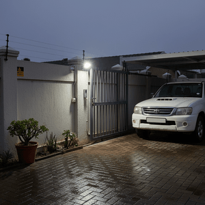 Wall-mounted floodlight above a metal gate lights a wet driveway and white SUV under a carport.