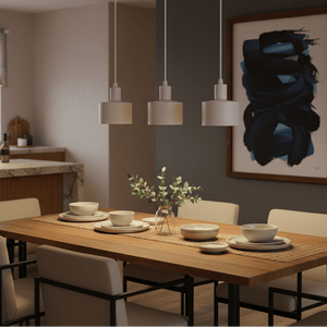 Three white metal pendant lights hang over a wooden dining table set with dishes and a vase of greenery.