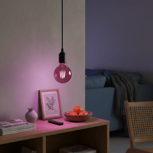 A pink filament bulb hangs above a wooden table with a bowl of oranges and a framed picture.