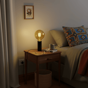 A bedside table with a decorative filament bulb in a lamp, surrounded by books and a mug.