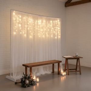 Twinkling icicle lights draped behind a white curtain, illuminating a wooden bench and table with candles.
