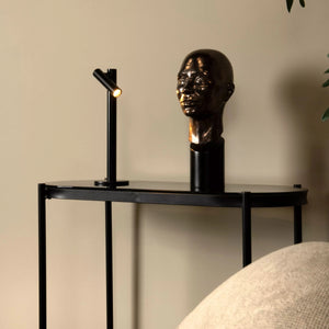 Decorative table with a bronze head sculpture and black lamp against a beige wall.