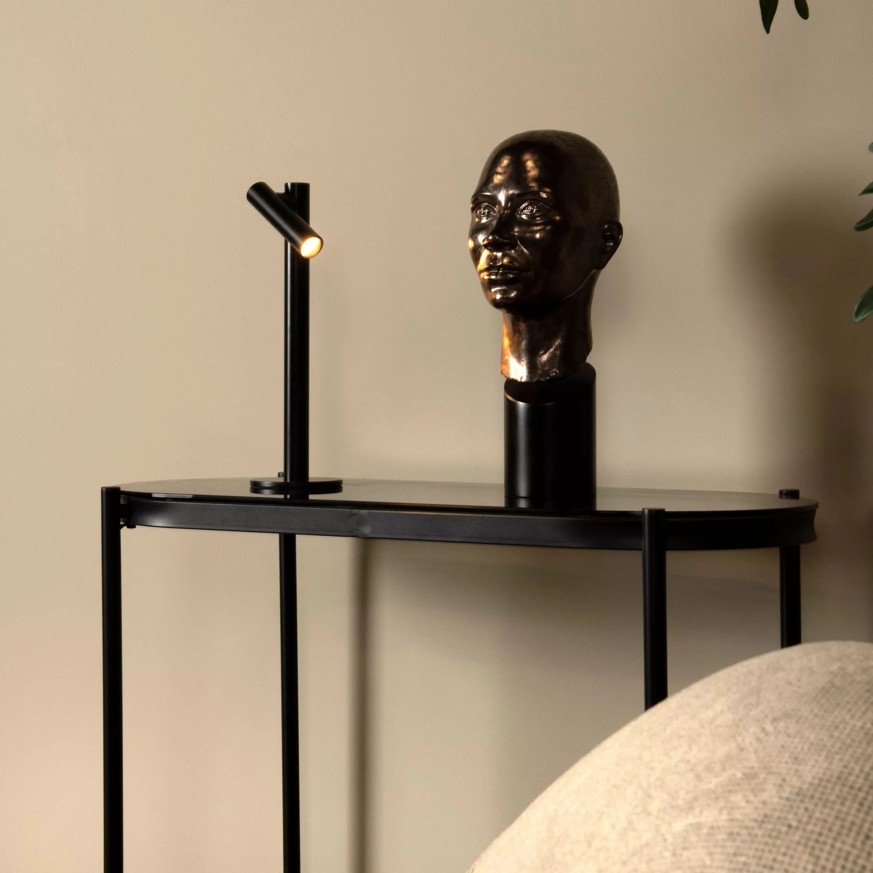 Decorative table with a bronze head sculpture and black lamp against a beige wall.