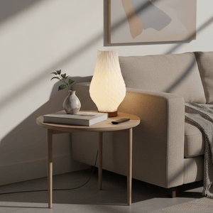 A modern 3D-printed table lamp on a wooden side table next to a cozy sofa, with a small vase and book.
