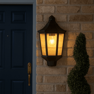 Black outdoor half lantern wall light illuminating a brick wall beside a blue door, with a small shrub nearby.