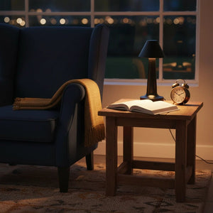 A cozy scene featuring a black rechargeable desk lamp on a wooden side table next to a blue armchair, with a book and alarm