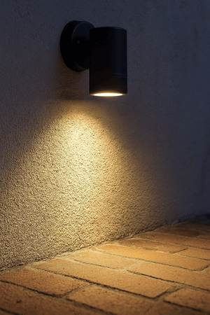Black cylindrical outdoor wall light mounted on a textured wall, casting warm downward light onto brick paving