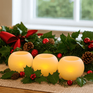 Three round LED candles glowing softly, surrounded by festive greenery and ornaments on a table.