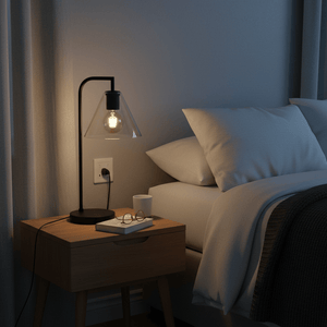A black table lamp with a clear glass shade sits on a wooden nightstand beside a bed with white pillows and a gray blanket.