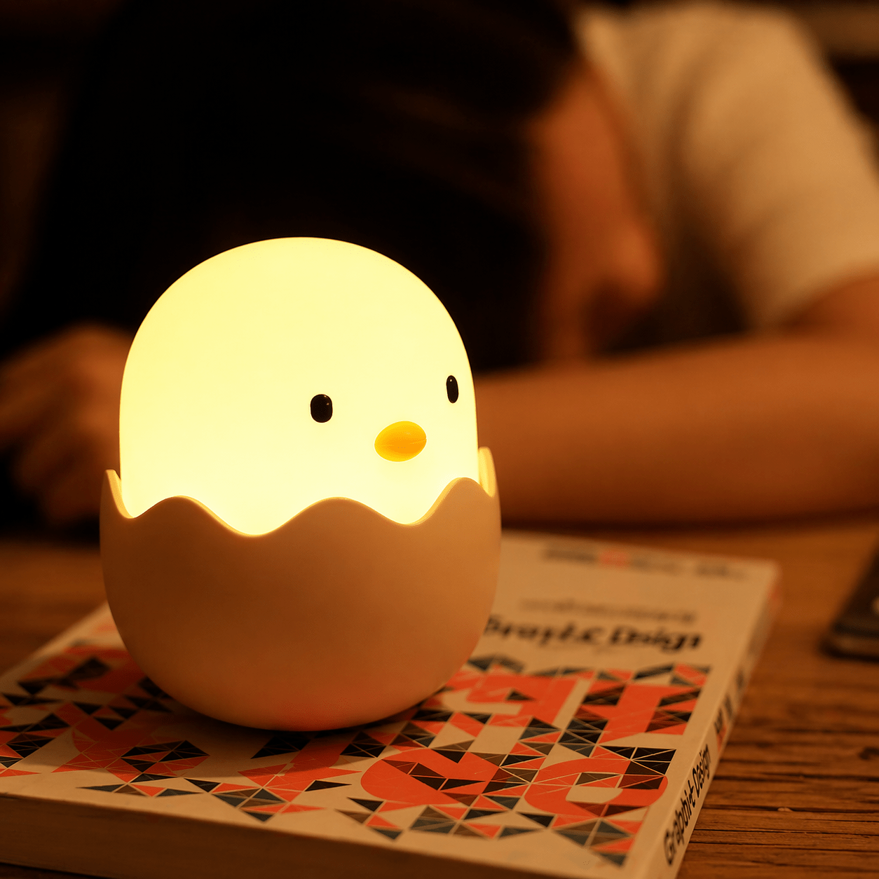 Glowing egg chick night light on a patterned book, with a sleeping child blurred in the background.