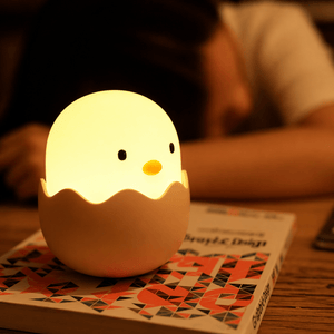 Glowing egg chick night light on a patterned book, with a sleeping child blurred in the background.