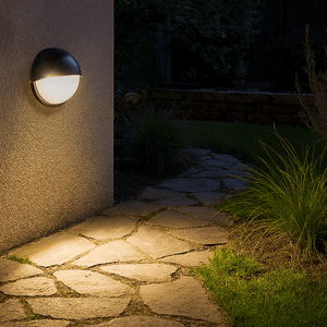 Black LED bulkhead light mounted on a textured wall, illuminating a stone pathway and surrounding grass.
