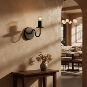 Harry Black Indoor Wall Light mounted on a textured wall, casting shadows, with a vase of dried flowers on a wooden table.