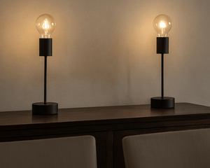 Two table lamps with illuminated bulbs on a wooden surface against a plain wall.