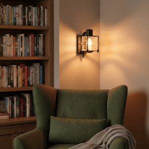 Matt black Torbay wall lamp illuminating a cozy reading nook with a green armchair and bookshelves.