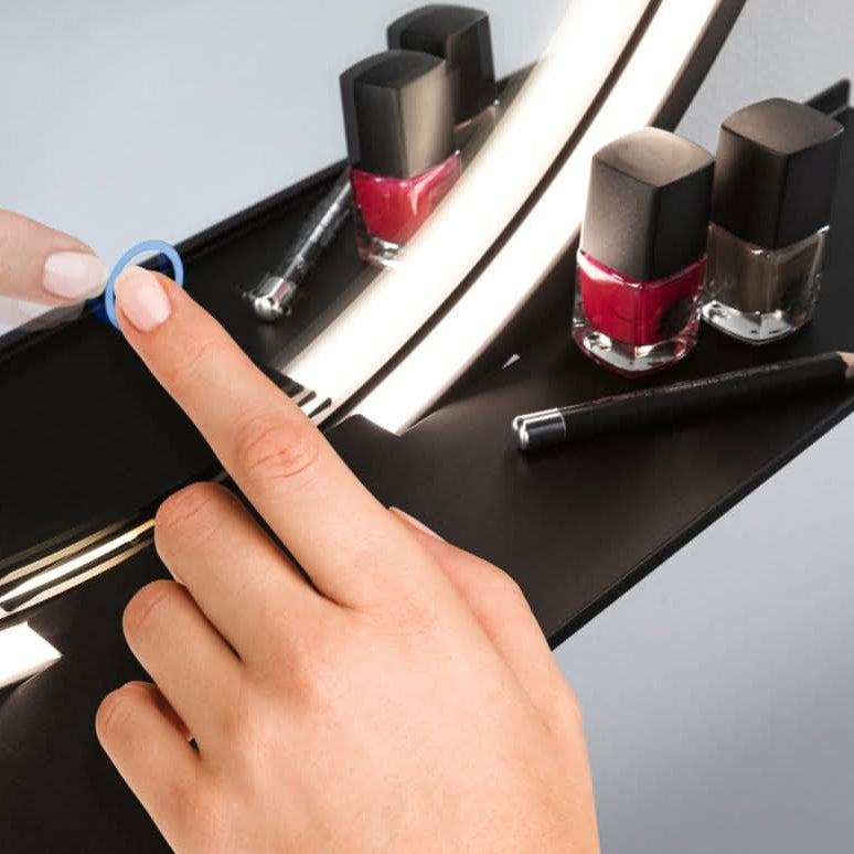 Close-up of a hand pointing at a blue ring on a black LED mirror, with nail polish bottles nearby.