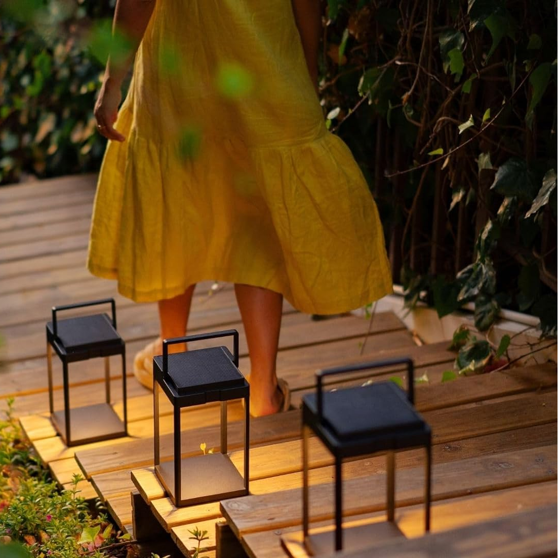Person in a yellow dress standing on a wooden deck with solar lamps, surrounded by greenery.