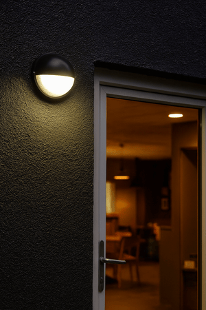 Black eyelid LED bulkhead light mounted on a dark textured wall beside a glass door.
