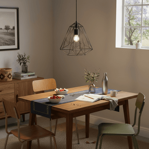 Black metal pendant light hanging above a wooden dining table set with fruit and a book.