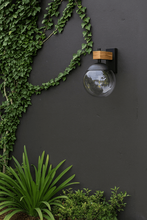 Bufalata Outdoor Wall Light mounted on a dark wall, surrounded by lush green plants.