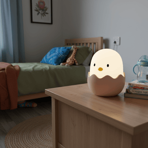 Chick-shaped egg night light glowing on a wooden bedside table in a child's bedroom with bed, books, and cup.
