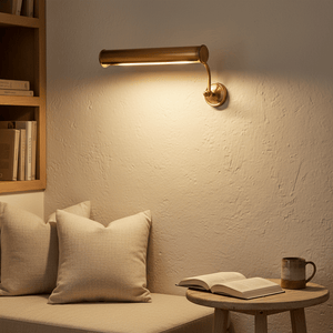 Solid brass wall lamp illuminating a cozy reading nook with pillows and a book on a table.