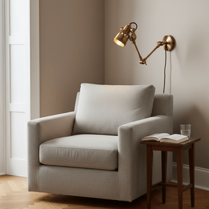 Indigo Swing Wall Lamp in antique brass mounted beside a cozy armchair with a side table and book.