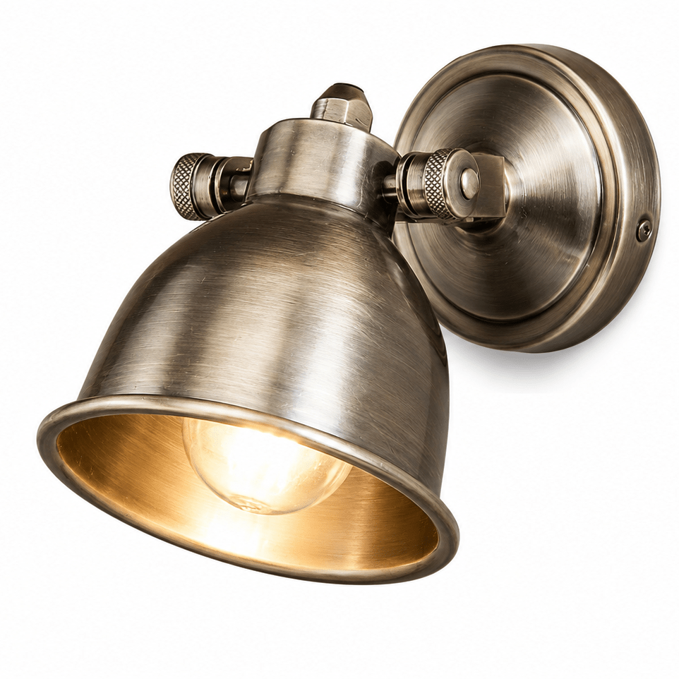 A wall-mounted industrial-style sconce with a dome-shaped shade and circular wall plate, shown in a brushed brass or bronze finish with the light turned on, against a plain white background.