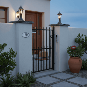 Black outdoor pillar lantern illuminating a garden entrance beside a gate, enhancing the pathway ambiance.