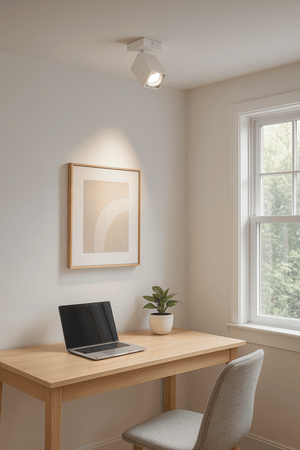 Square white spotlight mounted on ceiling, illuminating a minimalist workspace with a laptop and plant.