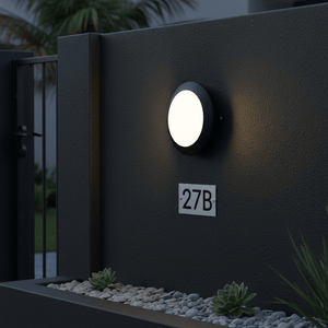 Outdoor wall light mounted on a dark wall, illuminating a pathway with decorative stones and a house number.