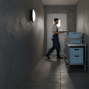 A man carrying a tray in a dimly lit hallway with a black bulkhead light mounted on the wall.