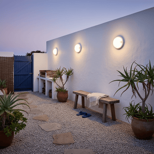 Three white bulkhead lights illuminate a courtyard with potted plants and a wooden bench.
