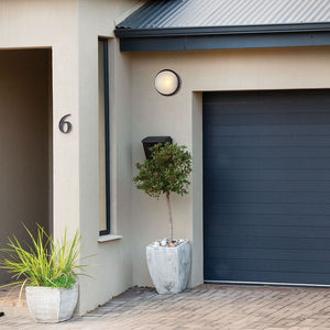 Large round black bulkhead light mounted on an outdoor wall beside a garage, with potted plants in the foreground.
