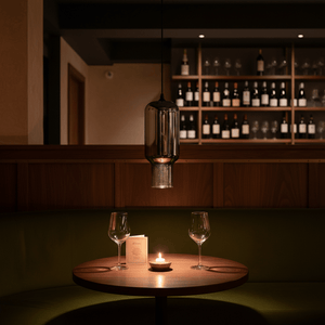 A black smoke glass pendant light hangs above a round wooden table with two wine glasses and a lit candle.