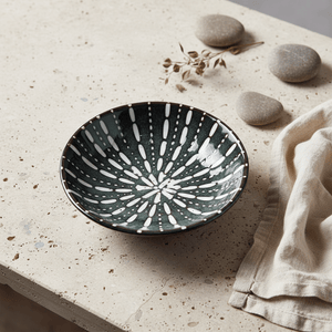 Ceramic flat bowl with a sea green sea urchin design on a textured surface, surrounded by pebbles and fabric.