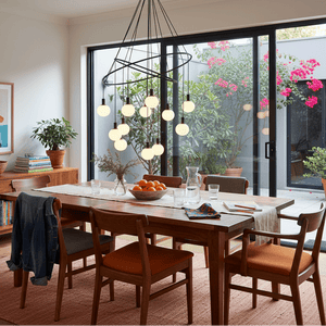 Cosmo 12 Light Chandelier hangs above a wooden dining table, surrounded by chairs and greenery.
