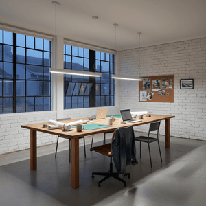 A modern office space featuring a wooden table with green papers, laptops, and coffee cups, illuminated by suspended linear