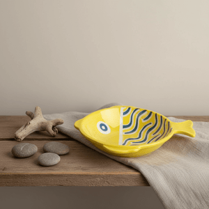 Ceramic fish-shaped bowl in yellow with blue line scales, placed on a wooden surface with pebbles and driftwood.