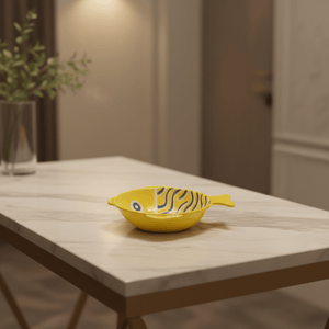 Ceramic fish-shaped bowl in yellow with blue line scales on a marble table.