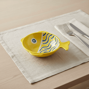 Ceramic fish-shaped bowl in yellow with blue line scales on a beige placemat, accompanied by cutlery.