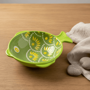 Ceramic fish-shaped bowl in green with yellow dot scales, placed on a wooden surface beside pebbles and fabric.