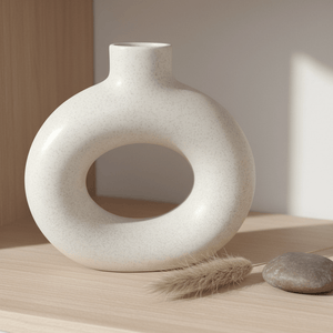 Small white ceramic vase in a hollow doughnut shape on a wooden shelf, accompanied by a dried flower and a smooth stone.
