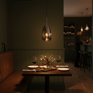 Smokey glass and chrome pendant light over a wooden dining table with plates, wine glasses, and cheese, bar area behind.