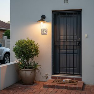 Black metal and glass outdoor wall light illuminating a front entrance with a potted plant and house number.