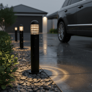 Montague 600 Bollard Light illuminating a wet driveway, surrounded by pebbles and greenery, with rain falling.